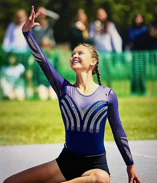 A gymnast in a sparkling blue leotard smiles and raises one arm gracefully during an outdoor performance.