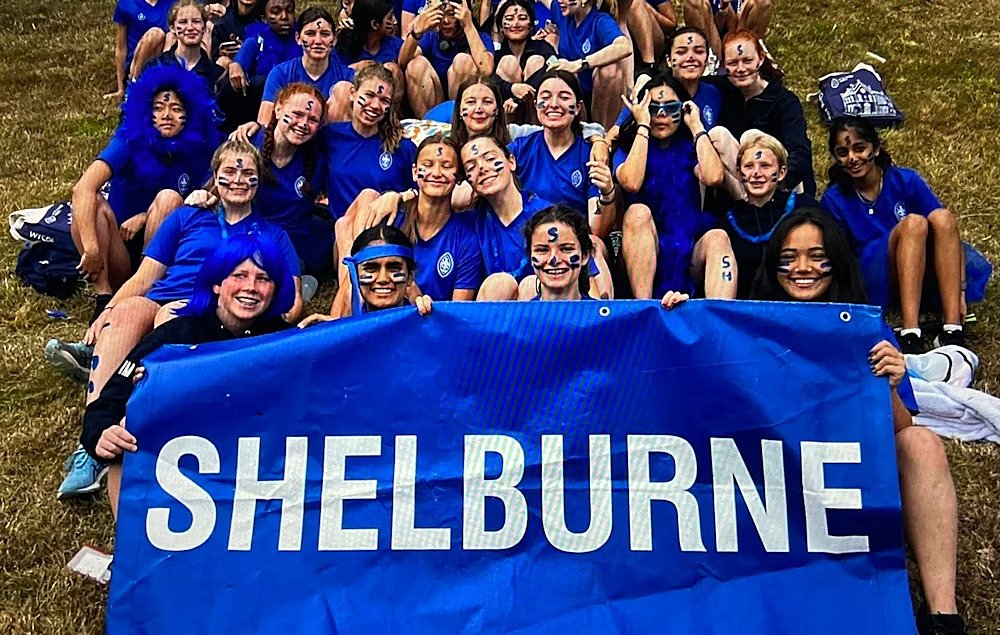 A large group of students wearing blue outfits and face paint hold a banner reading “Shelburne” and smile together.