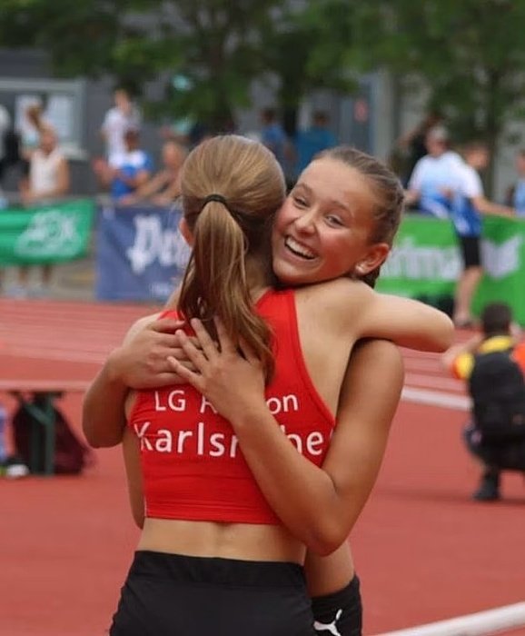 Two young athletes in red sports outfits embrace each other joyfully after a race on a running track.
