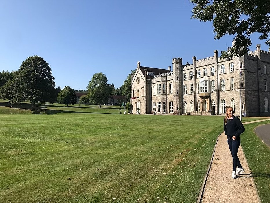 A student walks along a path in front of a large, historic school building surrounded by a green lawn on a sunny day.