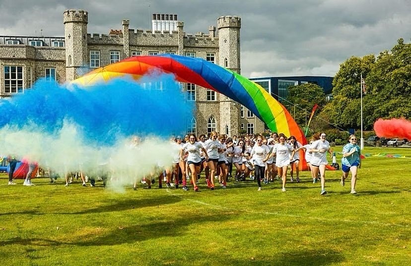 A group of runners start a colorful fun run on a school field with blue and yellow powder clouds and a rainbow arch.