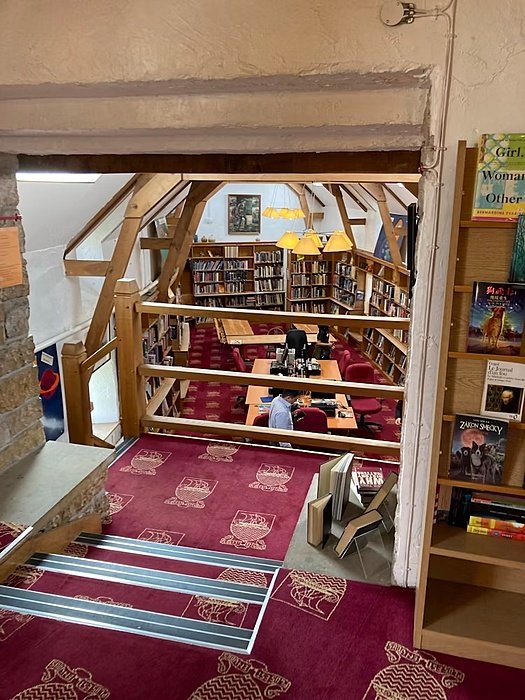 A cozy library with wooden beams, red carpet, and bookshelves filled with books, where students study at tables.