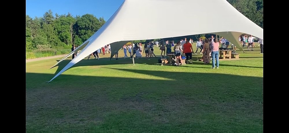 People gather under a large white canopy tent on a sunny day, socializing on a green lawn surrounded by trees.