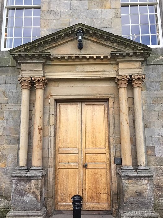 Large wooden exterior door with two pillars on each side and a pointed overhanging roof, set in a tall grey stone wall