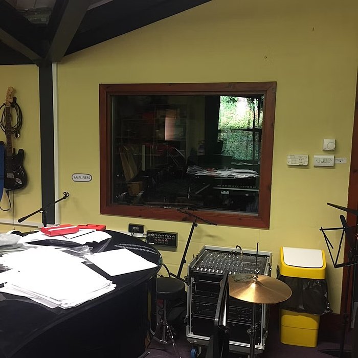 A busy room with drums and mix deck, a big window on the wall towards another music room