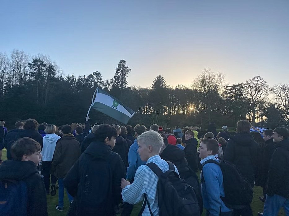 A group of students gather outdoors at sunset, one holding a green and white flag, surrounded by trees.