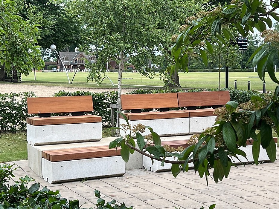 Modern outdoor benches with wooden seats and concrete bases surrounded by greenery, with a school field and pavilion in the background.