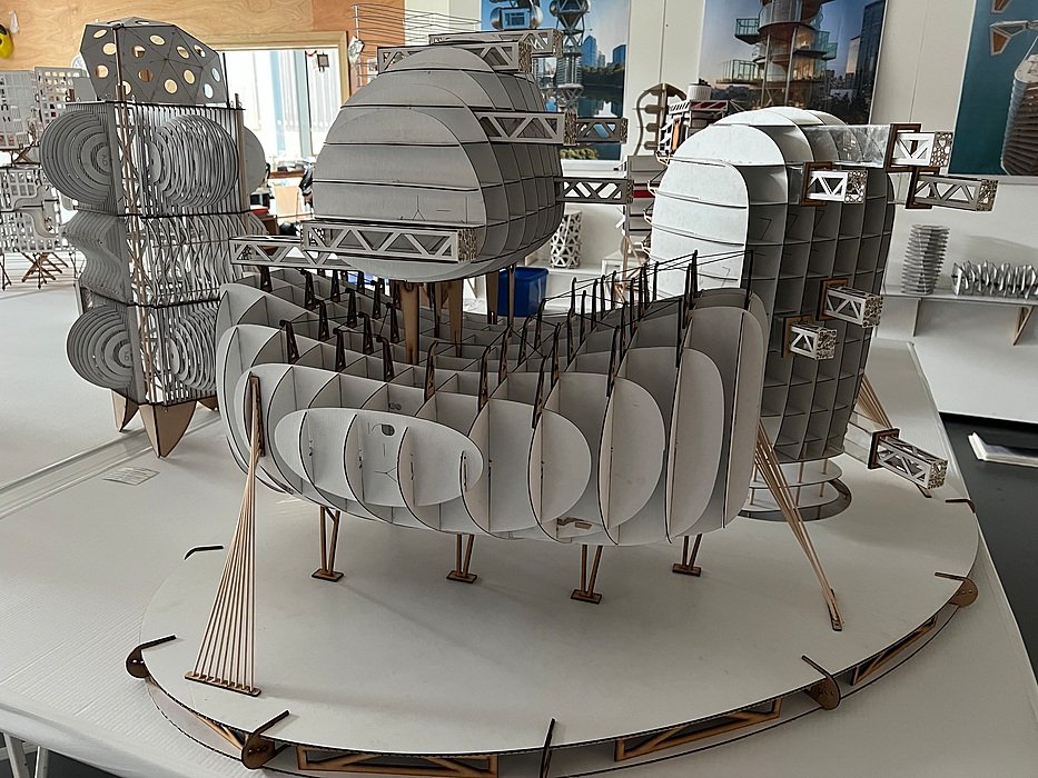 : Architectural scale models made from layered white card and wood, displayed on tables in a studio.