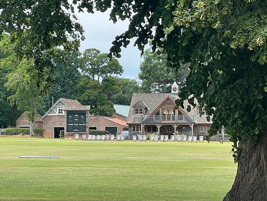 A cricket pavilion with a clock and scoreboard, surrounded by trees and open playing fields.