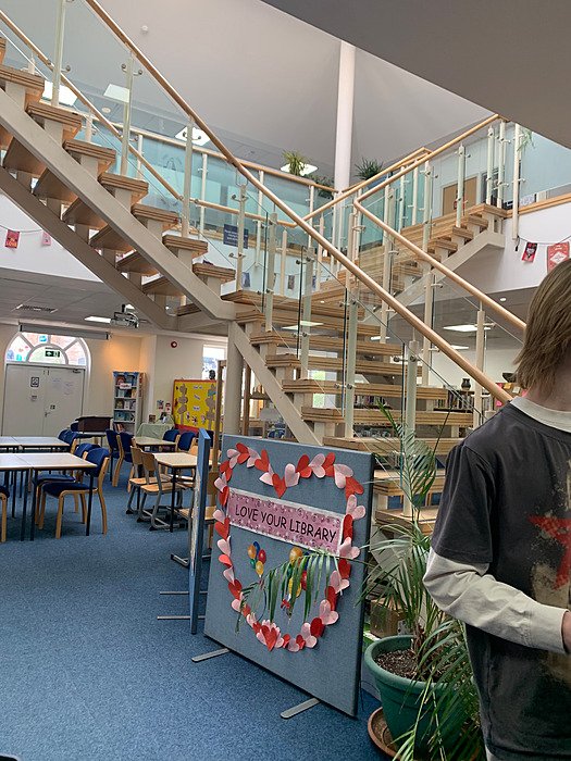 Library: A wide wooden staircase with glass balustrades crosses a bright room with tables, chairs and colourful posters.