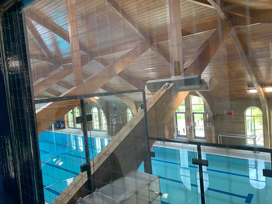 View through a glass wall onto an indoor swimming pool with blue water and a high timber roof.