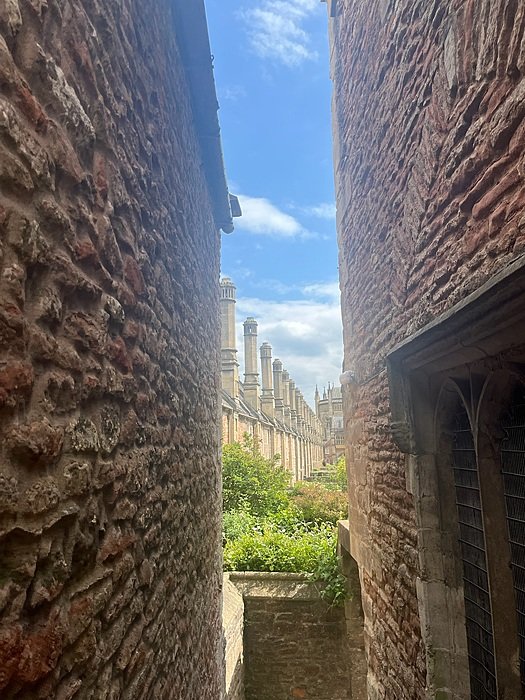 View through a narrow stone alleyway revealing the tops of ornate historic buildings and chimneys under a blue sky.