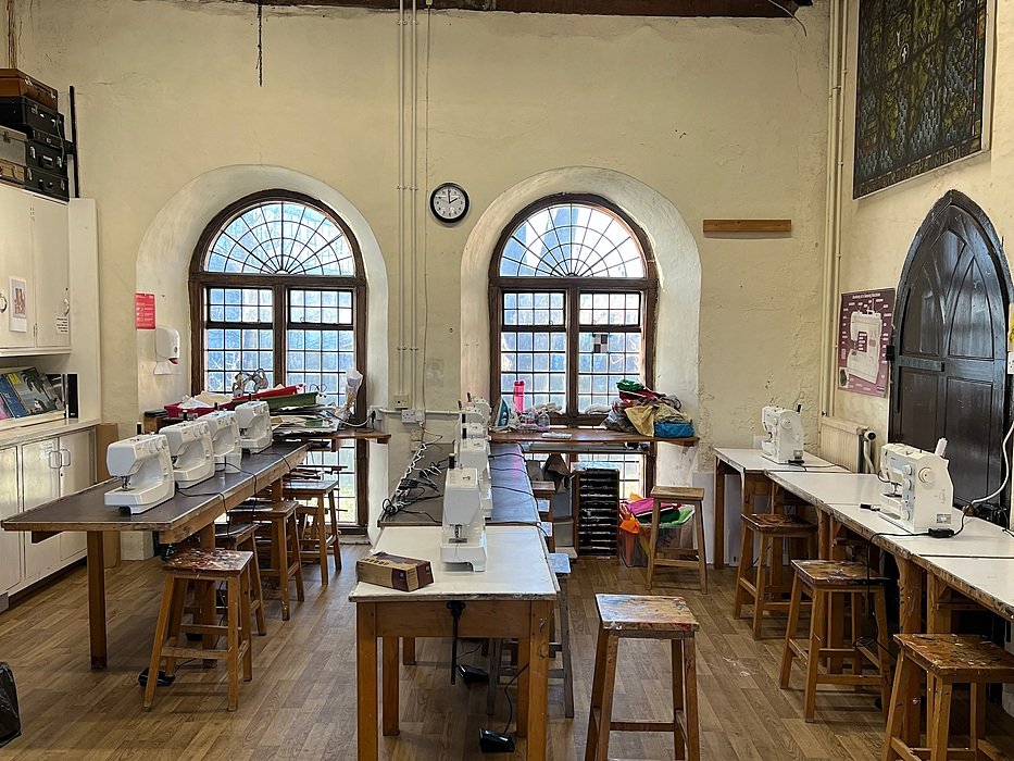 Classroom with sewing machines lined up on wooden tables beneath two arched windows in a historic building.
