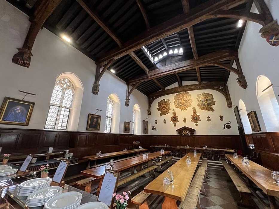 Historic dining hall with wooden beams, long tables, and coats of arms painted on the far wall.