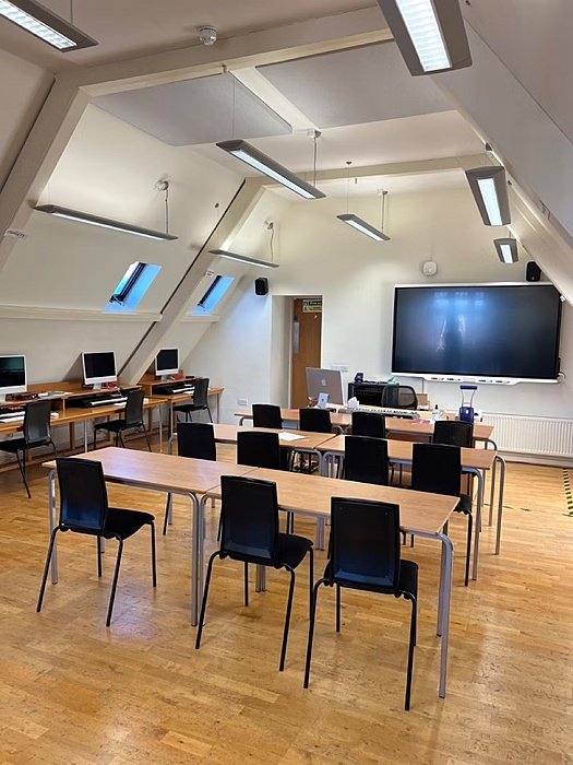 A classroom with rows of chairs and benches, computer desks on the side and a big screen behind the teacher's desk