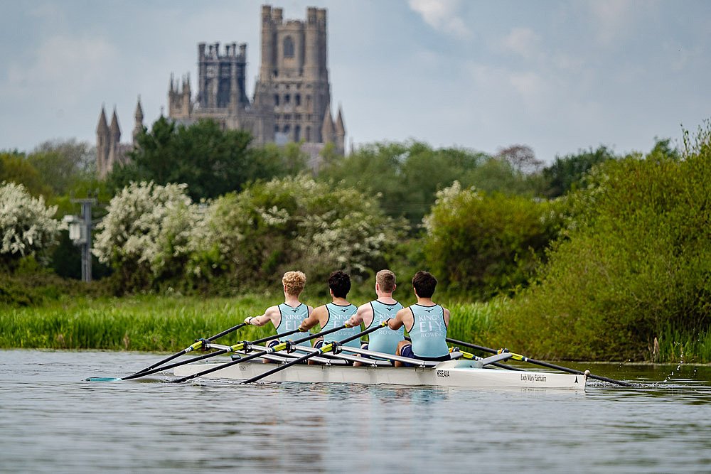 Four boys in a rowing boat on the water with a beautiful old School building in the background