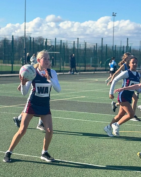 A girl is playing netball on an outdoor court while other players run in the background. She is wearing a school sports uniform with the letters “WA” on her bib.