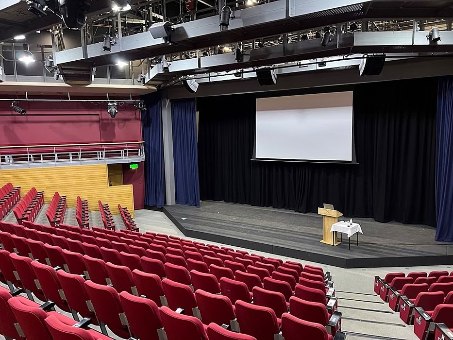 Red rows of seats for the audience facing the black stage with a white screen, a podium, and a table