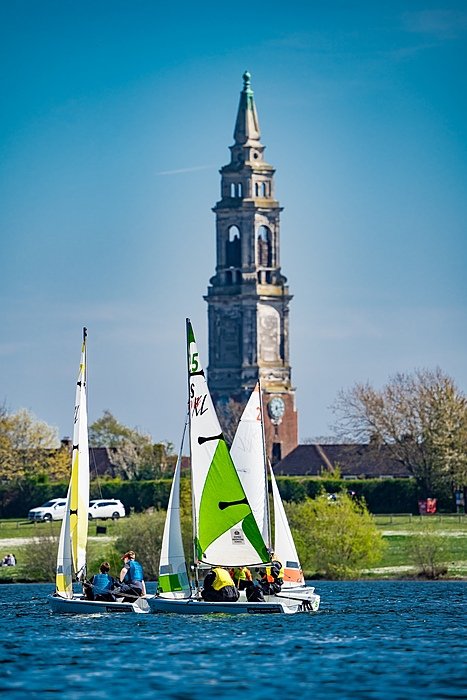 Pupils sailing small boats on a calm lake with the school’s tall clock tower rising in the background.