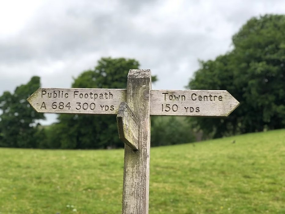 a wooden signpost in front of a green meadow with trees giving directions to the town centre and to a footpath
