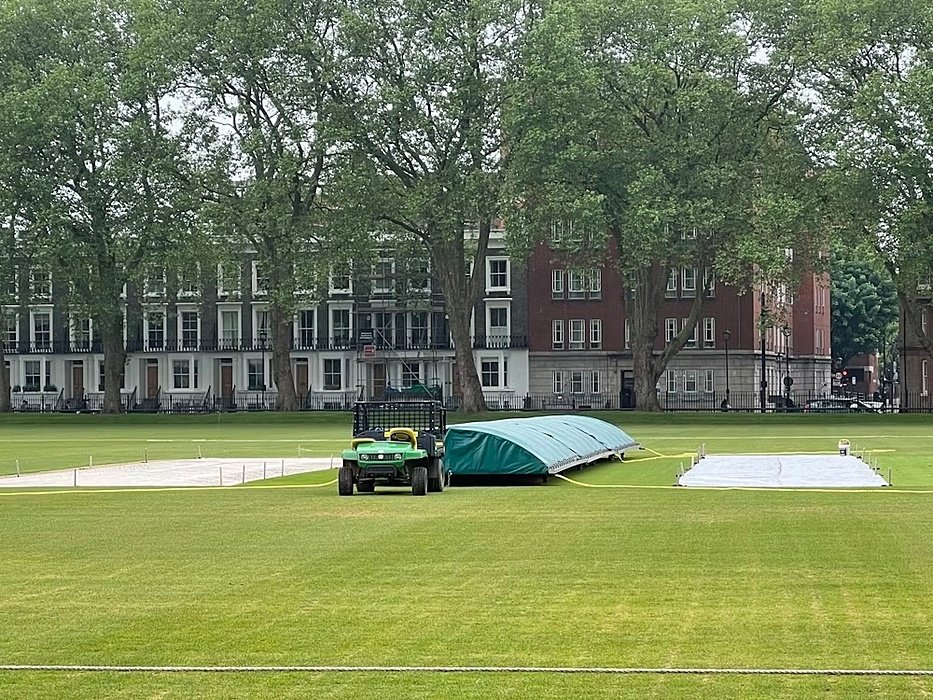 Cricket ground with covers on the pitch, surrounded by tall trees and Georgian-style buildings.