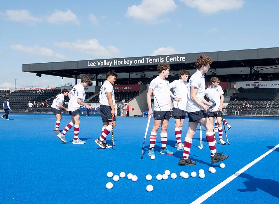 Group of boys in white and blue hockey uniforms standing on a blue hockey field with white balls, at the Lee Valley Hockey and Tennis Centre.