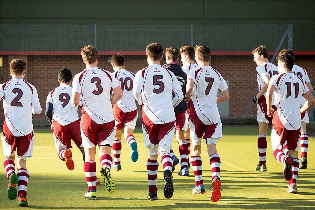 Group of boys in white and maroon sports uniforms running together on a hockey field during training.