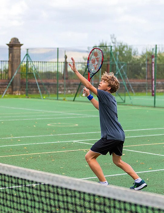 A student playing tennis outdoors, preparing to serve on a green court surrounded by fencing.
