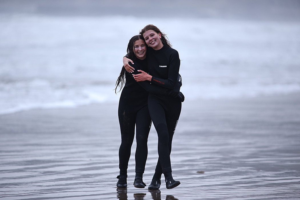 Two friends in wetsuits hugging and smiling on a beach after surfing.