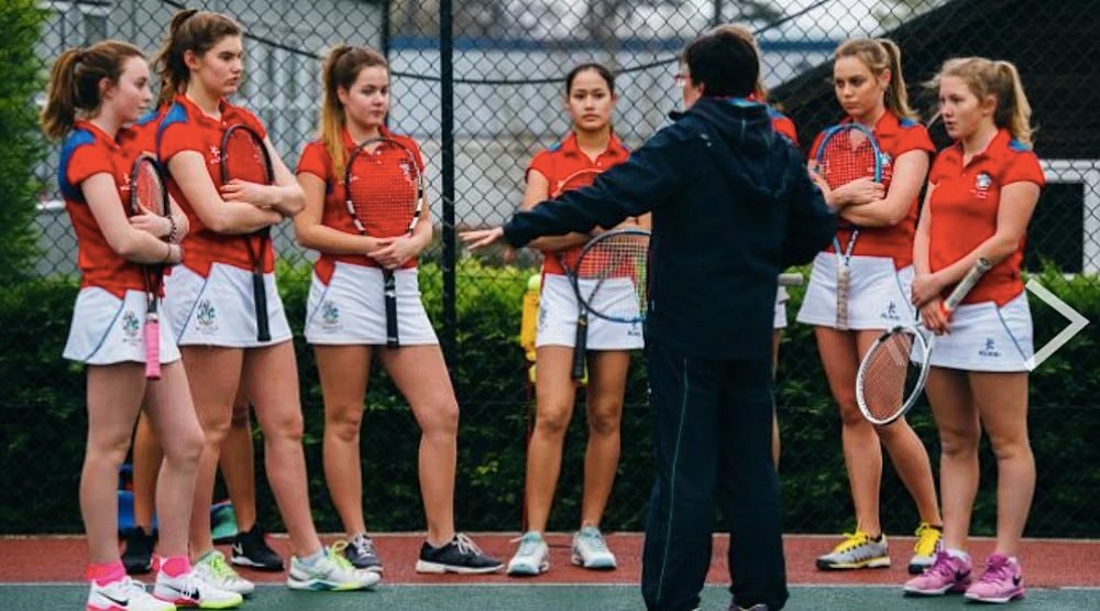 Coach giving instructions to a girls’ tennis team gathered on the court.