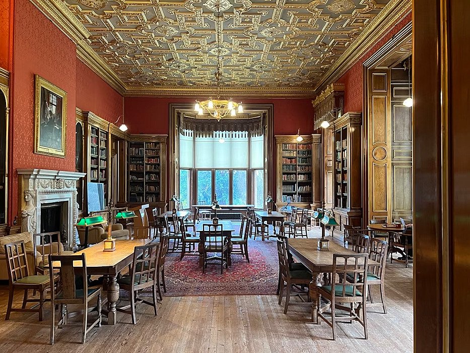 A grand library with wooden furniture, red walls, a patterned ceiling, and shelves full of books.