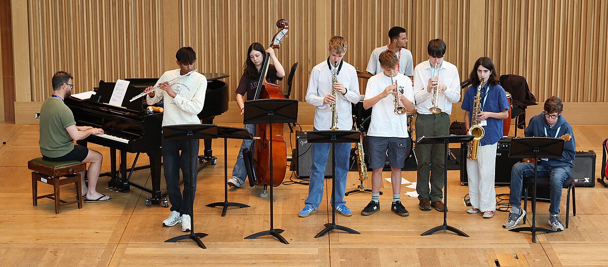 A group of students playing instruments including trumpet, saxophone, and piano during a music rehearsal on a wooden stage.