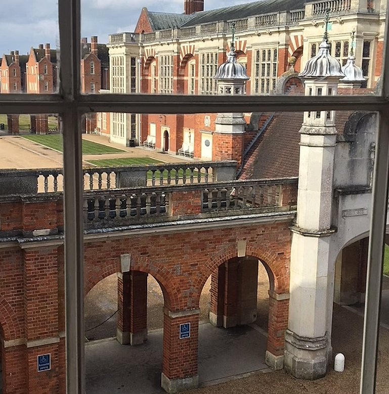 View through a large window of a grand courtyard surrounded by ornate red-brick and stone buildings.
