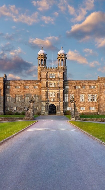A panoramic view of a grand stone school building with towers and a long driveway, surrounded by lawns and gardens.
