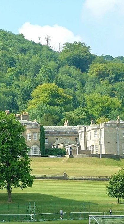 A grand country house surrounded by open lawns and woodland, set at the base of a green hillside.