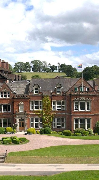 A large red-brick school building surrounded by lawns and trees, with a grand entrance and countryside hills in the background.