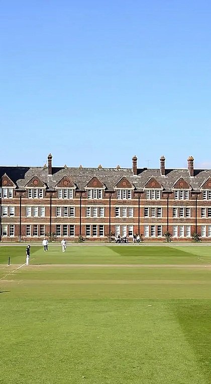 Students play cricket on a large green field in front of a grand red-brick school building with a clock tower.