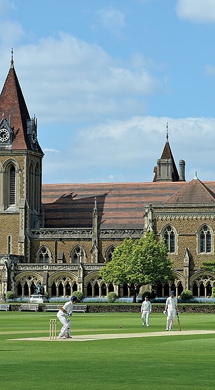 A cricket match taking place on a manicured green field in front of a grand historic school building with towers and arched windows.
