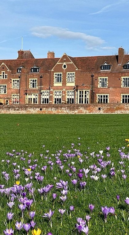 A historic red-brick school building with a large lawn and colourful spring flowers blooming in the foreground.