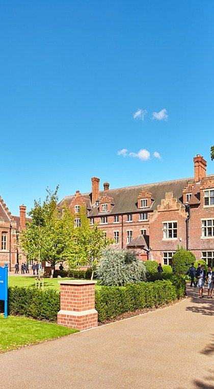Wide-angle shot of a school campus with green lawn and pupils on foreground and red-brick school buildings in back