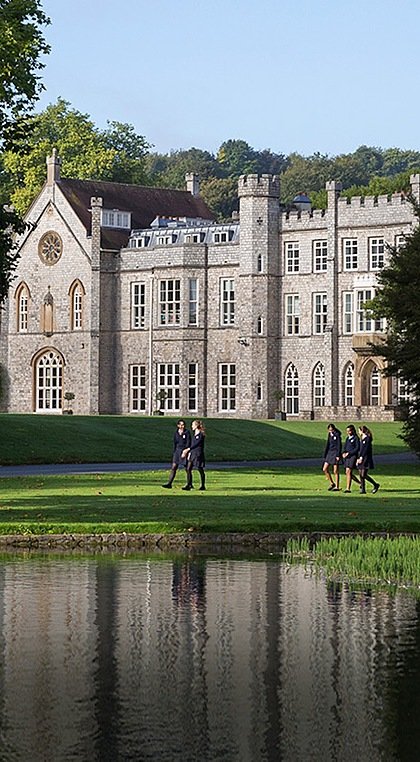 An imposing, grey manor house building with a lake and trees in the foreground with girls going for a walk