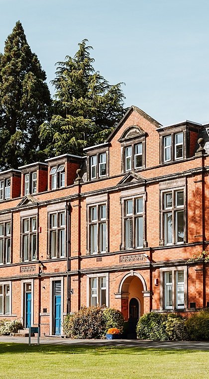 Historic red-brick school building with large windows and a clock tower surrounded by trees.