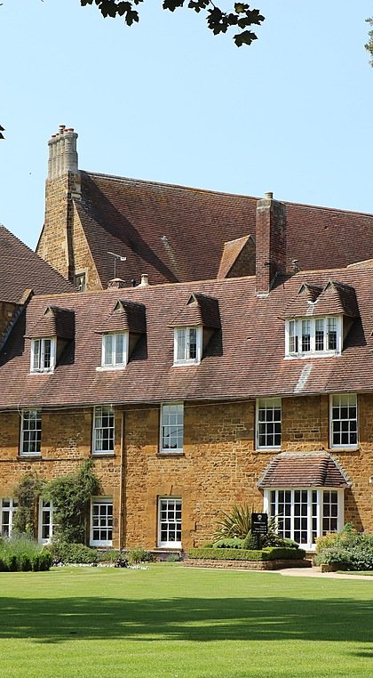 A historic brick building with gabled roofs surrounded by trees and a well-kept lawn.