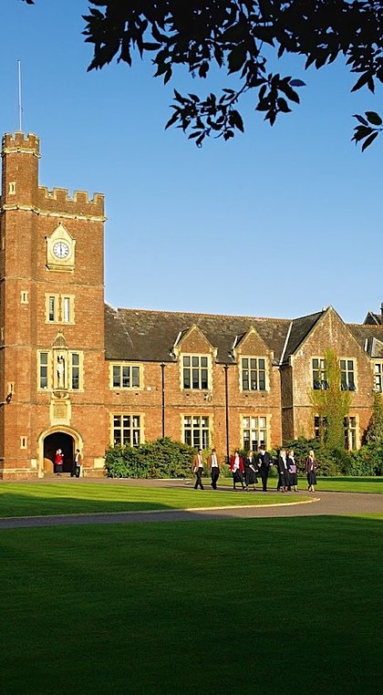 A large red-brick school building with a central clock tower and chapel, surrounded by neatly cut lawns and students walking in the courtyard.