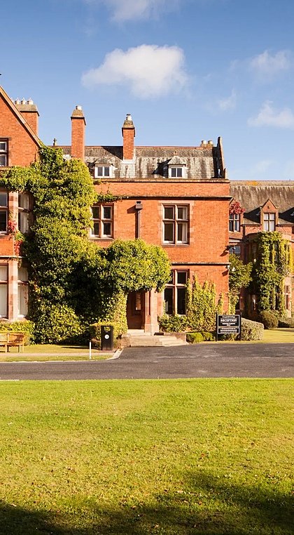 A red-brick school building with ivy-covered walls and large windows, set around a courtyard with green lawns.