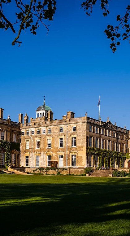 A stately country house with ivy-covered walls and a domed roof, surrounded by open green lawns and framed by tree branches.