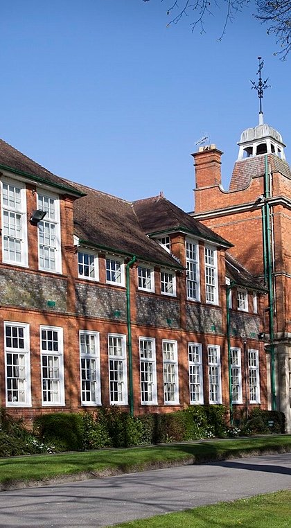 A long red-brick school building with tall windows and a clock tower, lined with trees and neatly kept lawns.