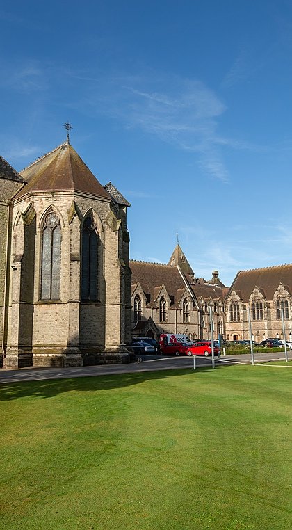 A grand stone chapel and adjoining school buildings set beside a neatly kept lawn under a clear blue sky.