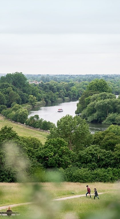 Scenic river view framed by green trees and open fields with a footpath and two people walking.