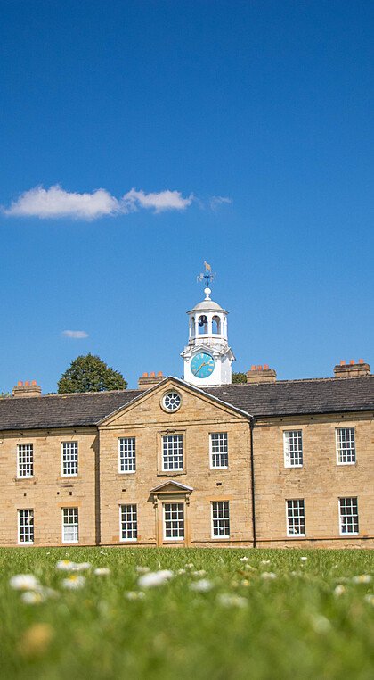 A historic sandstone school building with a clock tower and cupola, set against a bright blue sky and surrounded by a well-kept lawn.